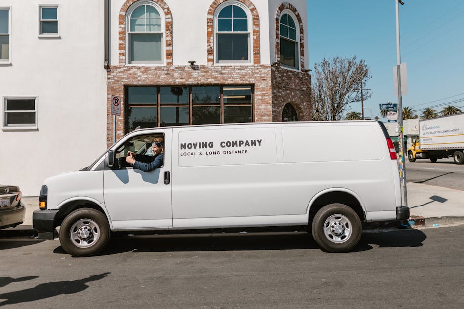 A smiling man with a full beard and an afro hairstyle stands outside next to a white moving van with the company logo 'MOVING COMPANY LOCAL & LONG DISTANCE' visible on the side. The man is wearing a n