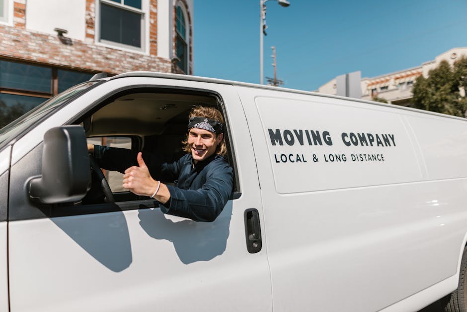A young male mover with long hair, wearing a patterned headband and a navy blue shirt, is sitting in the driver's seat of a white moving van operated by Man With a Van Parsons Green. The mover is smiling and giving a thumbs-up, indicating a positive attitude towards home relocation or furniture transport. The van's side panel displays the company name and the phrase 'Local & Long Distance,' suggesting comprehensive removals services. The background shows a clear blue sky, surrounding buildings with brick and modern facades, and some trees, indicating an urban environment typical of Parsons Green. Inside the van's open door, part of the interior can be seen, with cardboard boxes and packing materials visible, hinting at the packing and moving process. The scene captures the loading or unloading phase of a house removal, with the focus on professional logistics and transportation for residential relocations.