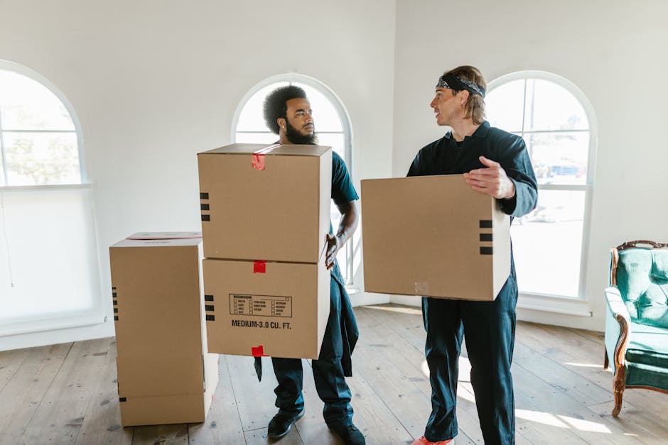 Two professional movers from Man With a Van Parsons Green in a well-lit room with wooden flooring and white walls, engaged in a home relocation process. The mover on the left, with an afro hairstyle and beard, is carrying a large cardboard box with a red tape and a printed label, and has a focused expression. The mover on the right, wearing a headband and casual dark clothing, is holding a medium-sized cardboard box and appears to be explaining or coordinating the loading process. Several other cardboard boxes are stacked nearby, some sealed with red tape and labeled for transport. The room features two large arched windows allowing natural light to fill the space, with a green upholstered armchair partially visible on the right side. The scene depicts the packing and furniture transport phase of a household move, highlighting the careful handling of packed items during loading, facilitated by using cardboard boxes, plastic wrap, lifting equipment, and a moving van possibly parked outside. Man With a Van Parsons Green's team ensures organized and professional packing and moving services.