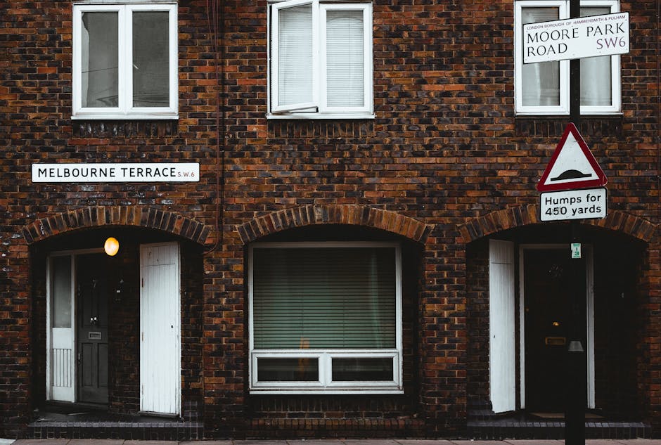 Photograph of a brick residential building on Melbourne Terrace, SW6, during a house removal process. The building has two windows on the upper floor with white frames and partially closed blinds. The