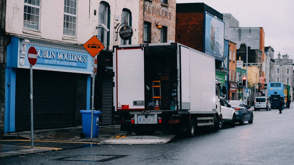 A white moving van with its rear doors open is parked on a street in Parsons Green, London, during the day. The van is positioned next to the sidewalk, with some cardboard boxes, plastic wrapping, and