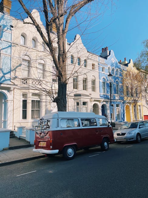A vintage red and white Volkswagen camper van is parked on a residential pavement in front of a row of colorful Victorian-style terraced houses, with large windows and ornate facades, under a clear blue sky. The van is positioned close to the curb, with a roof rack attached at the rear, possibly used for securing boxes or bags during a home relocation. Nearby, a silver car is also parked on the street, and a leafless tree extends its branches over the scene, indicating it may be late autumn or winter. The setting suggests a quiet suburban area suitable for house removals and furniture transport, with [COMPANY_NAME] often utilising such vans during the loading process. The image captures a typical moment in professional moving services, where packing and loading of household items into the vehicle take place in an organized manner, supporting efficient logistics for local or long-distance moves in the Parsons Green area.